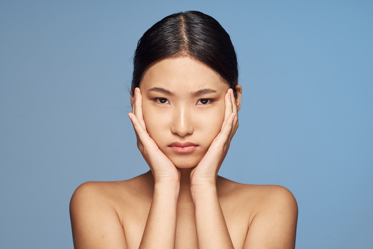 A sad Asian woman with bare shoulders touches her face, gray isolated background. 